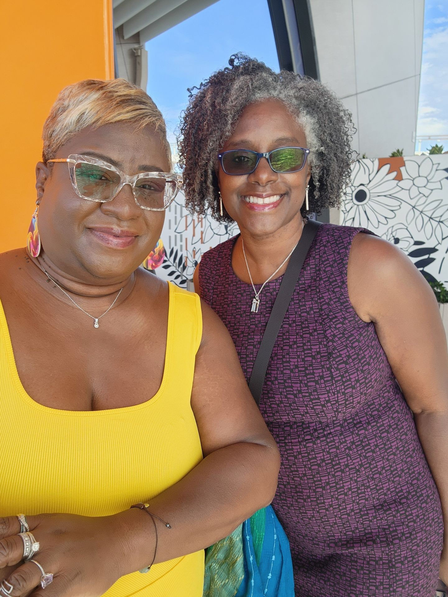 Two women smiling, standing together against a colorful and patterned wall background.