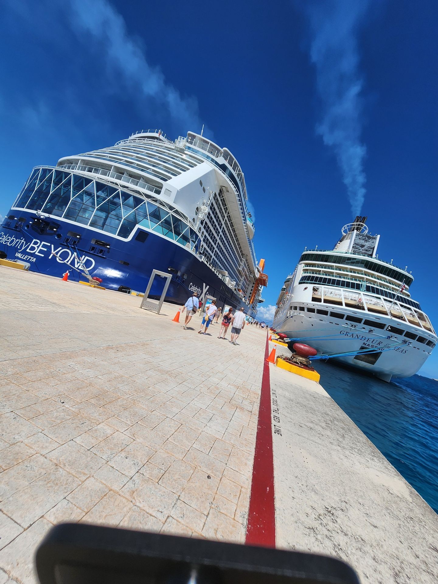 Two cruise ships docked at a sunny pier with people walking along the path.